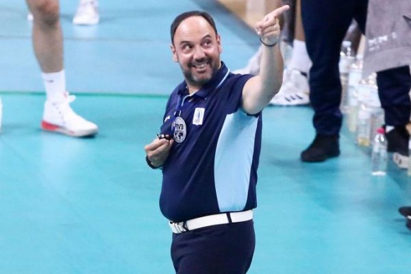 Volleyball coach on an indoor court, pointing and smiling while wearing a dark blue polo with light blue panels and a white belt.