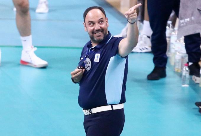637697330014598911 The Volley Lovers Volleyball coach on an indoor court, pointing and smiling while wearing a dark blue polo with light blue panels and a white belt.