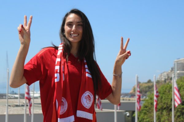 Woman in a red shirt and scarf, smiling and making peace signs with both hands outdoors on a sunny day.