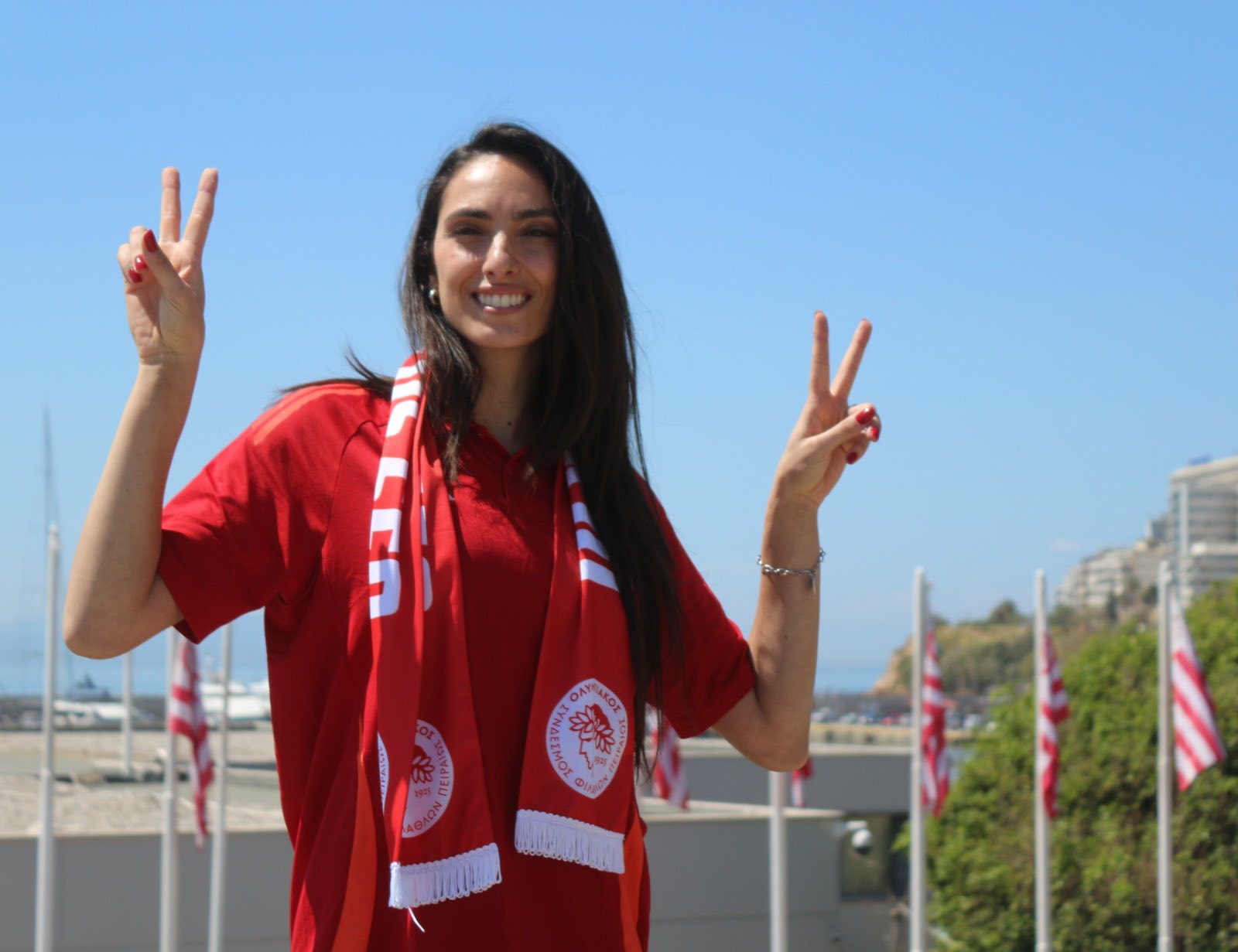70090794814066234414939825231036891299456421n The Volley Lovers Woman in a red shirt and scarf, smiling and making peace signs with both hands outdoors on a sunny day.