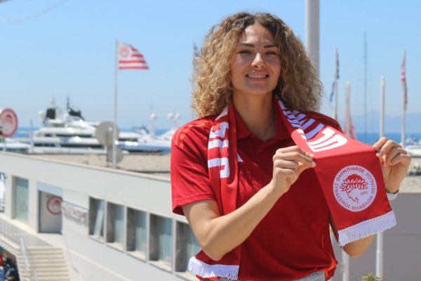 Woman in a red shirt proudly holds a red scarf with a circular emblem, standing at a sunny marina with yachts and flags in the background.