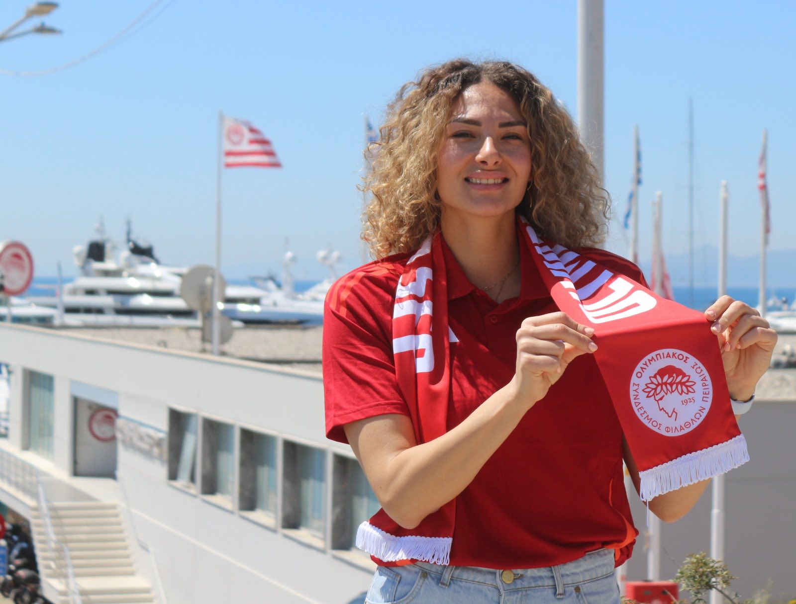 70363588314074921380737794487537629708866112n The Volley Lovers Woman in a red shirt proudly holds a red scarf with a circular emblem, standing at a sunny marina with yachts and flags in the background.