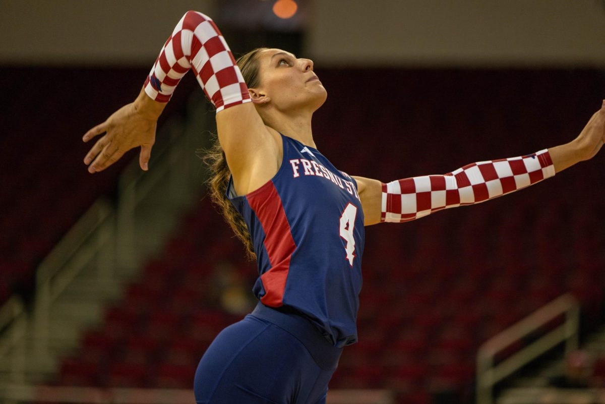 Female gymnast in a blue uniform with red side panels and red-and-white checkerboard sleeves, arms raised in a dramatic stretch inside a stadium or arena.