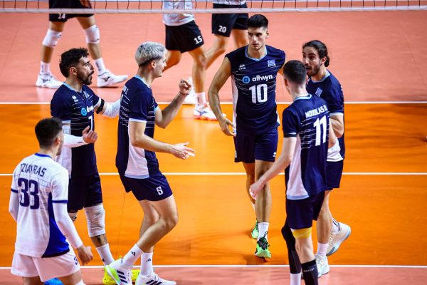 Six male volleyball players in navy and white uniforms celebrate on an orange indoor court near the net, giving each other high-fives.