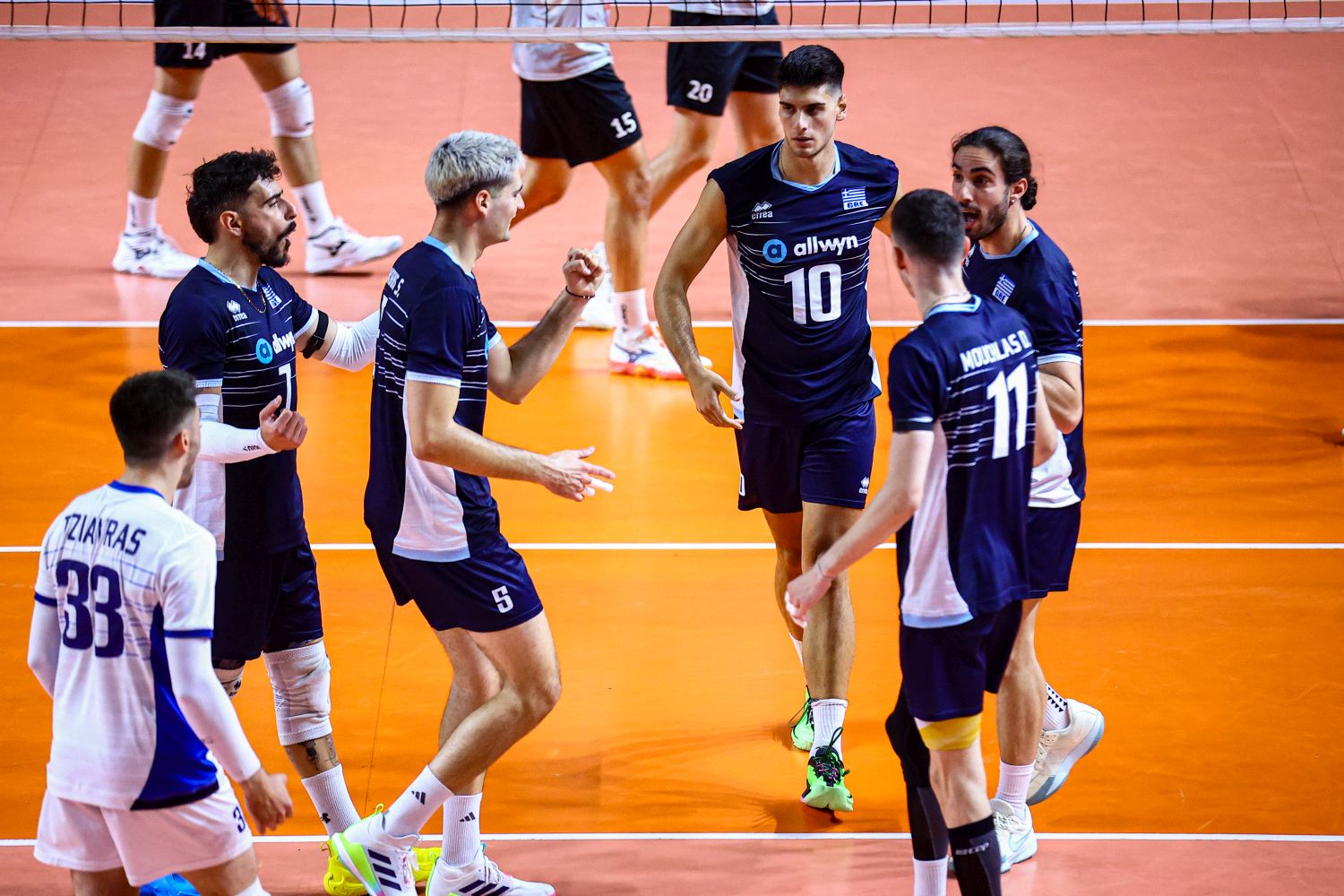 Six male volleyball players in navy and white uniforms celebrate on an orange indoor court near the net, giving each other high-fives.
