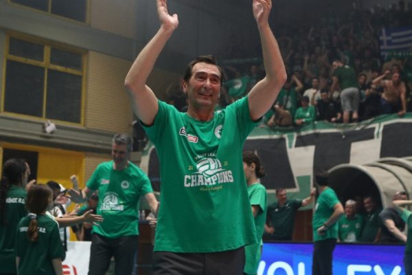Man in a green 'Champions' shirt raises his arms in celebration at an indoor volleyball arena with cheering fans in the background.