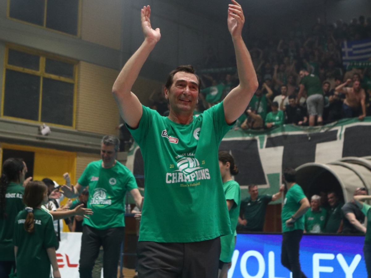 Man in a green 'Champions' shirt raises his arms in celebration at an indoor volleyball arena with cheering fans in the background.