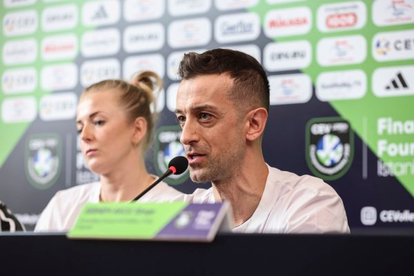 Man speaking into a microphone at a press conference, with a blurred woman in the background and a sponsor logo backdrop.