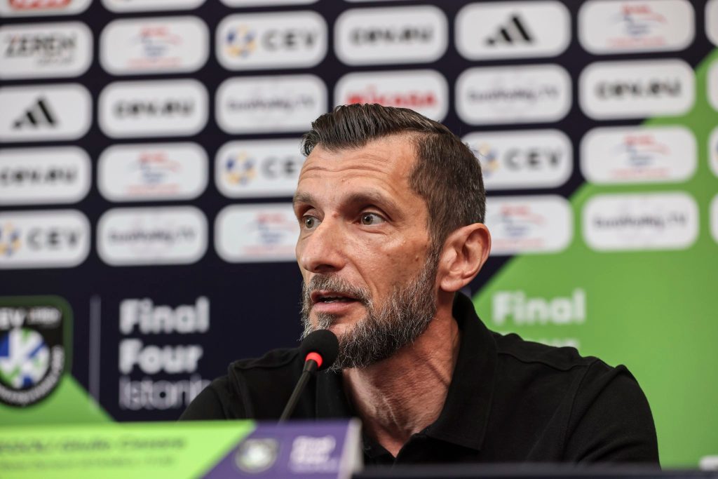 ecz1 The Volley Lovers Man with short dark hair and beard speaks into a microphone at a press conference backdrop of sponsor logos.
