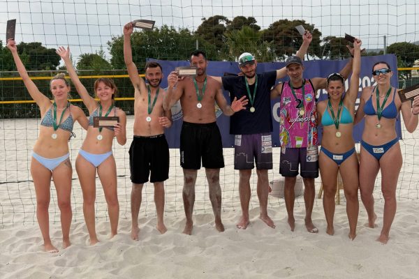 Nine beach volleyball players stand on sand behind a net, wearing medals and holding plaques in celebration photo.