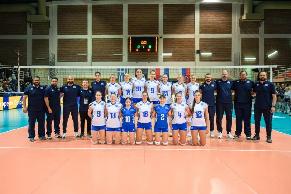 Group photo of a women's volleyball team and coaches on an indoor court, kneeling in front of the net with players in white and blue uniforms.