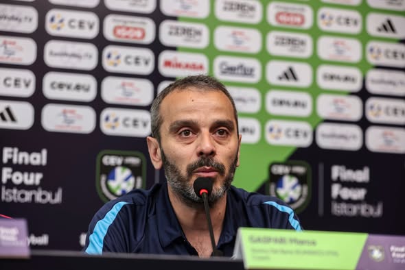 sca1 The Volley Lovers Bearded man at a press conference speaking into a microphone, with sponsor logos in the background.