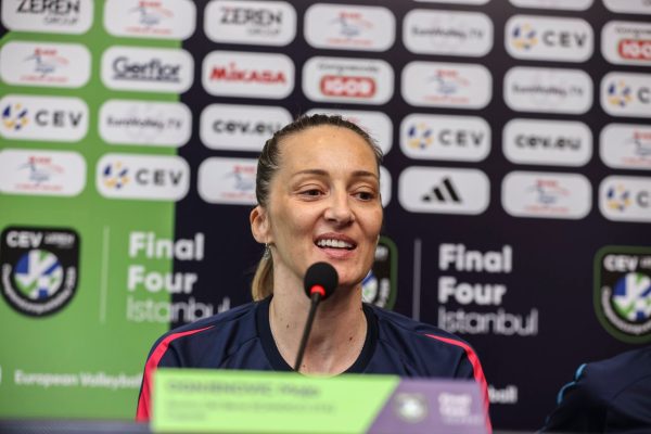 Smiling female athlete at a press conference speaking into a microphone, with a sponsor-filled backdrop and 'Final Four' graphics behind her.