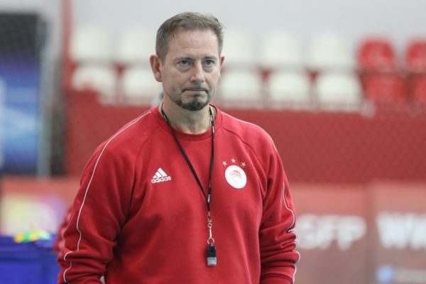Male coach in a red Adidas sweatshirt with a lanyard and whistle, standing in an indoor sports arena.