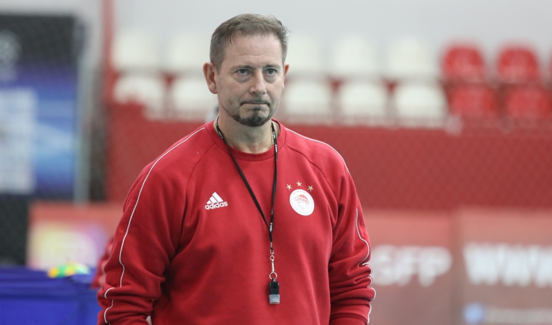Male coach in a red Adidas sweatshirt with a lanyard and whistle, standing in an indoor sports arena.