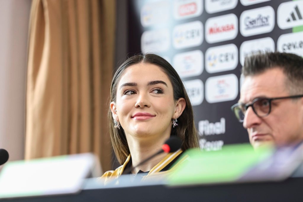 vak1 The Volley Lovers Close-up of a smiling woman with dark hair at a press conference, microphone in foreground and sponsor logos on a backdrop behind her.