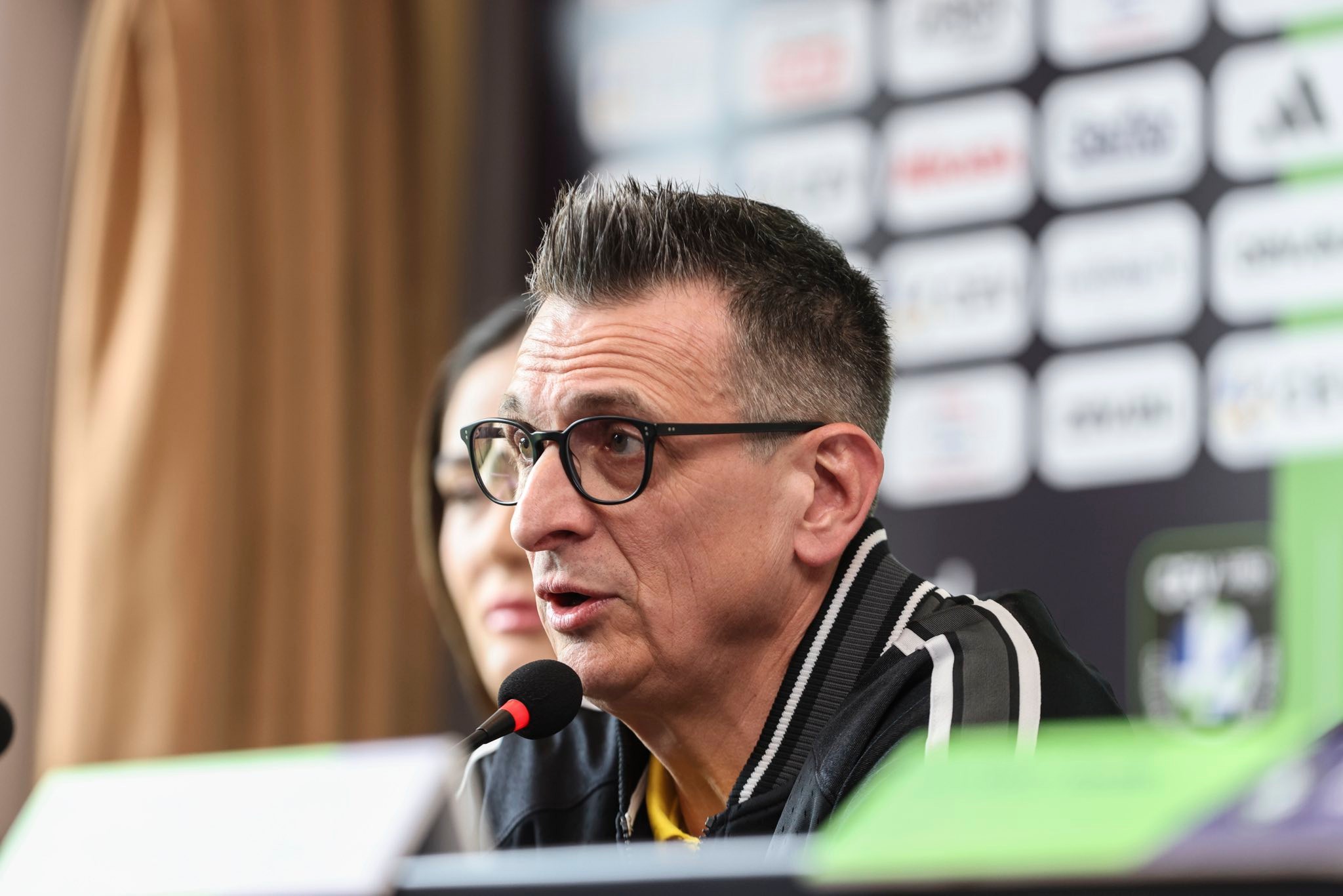 vak2 The Volley Lovers Man wearing glasses speaks into a microphone at a press conference with a blurred sponsor backdrop behind him.