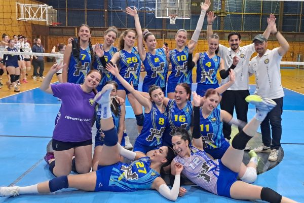 Group of female volleyball players in blue jerseys celebrate on an indoor court, arms raised and smiling.