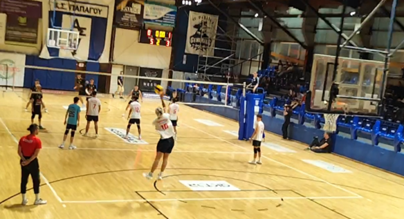 Indoor volleyball game in a gym with players in white jerseys; one player is jumping for a spike near net, coach or referee watching nearby.
