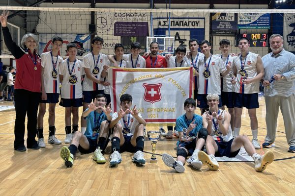 Youth volleyball team with medals posing in a gym, holding a white banner featuring a red shield and Greek text; trophy on the floor in front of them.