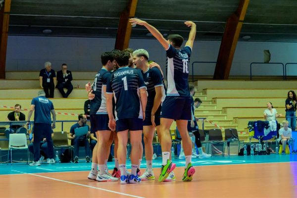A group of male volleyball players in blue uniforms huddle and celebrate on an indoor court.