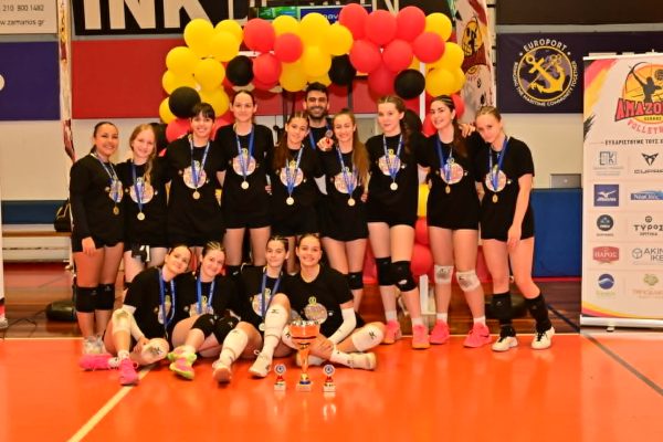 Team of young volleyball players in black uniforms pose with medals and trophies in a gym; colorful balloon arch behind them.