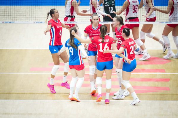 Group of volleyball players in red and blue uniforms celebrate together on a bright indoor court near the net, pink shoes visible.