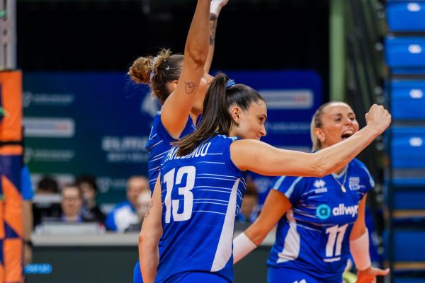 Volleyball players in blue uniforms celebrate on court, arms raised in victory, with a teammate wearing number 13 in front.