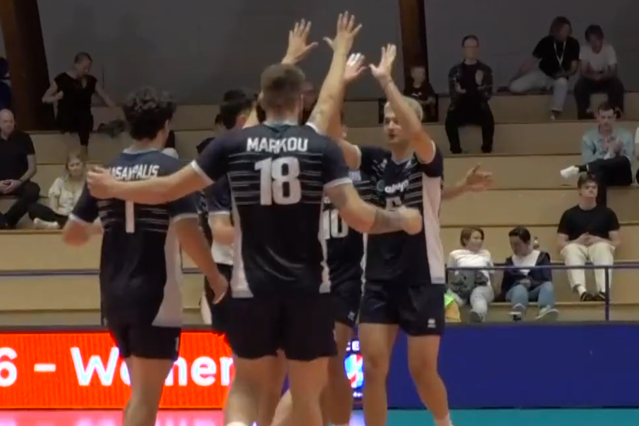 Volleyball players in dark uniforms high-fiving in a huddle on the court as teammates celebrate a point, with spectators in the stands behind them.
