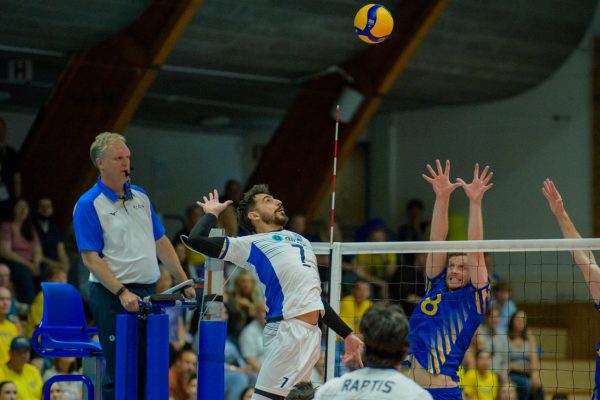 Volleyball spike moment: attacker in white-blue jumps to hit the ball over the net while blockers leap to defend and the referee watches from a blue chair nearby.