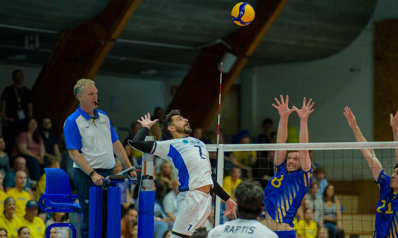 Volleyball spike moment: attacker in white-blue jumps to hit the ball over the net while blockers leap to defend and the referee watches from a blue chair nearby.