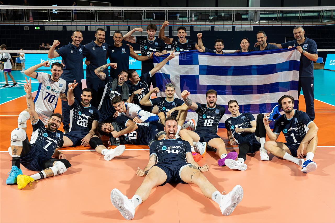 Male volleyball players celebrate on an indoor court, holding a large Greek flag and cheering in dark blue uniforms.