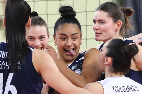 A group of female volleyball players in navy uniforms celebrate in a tight huddle, one player sticking her tongue out playfully in the center.