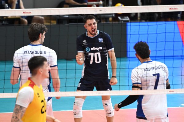 Male volleyball player in dark jersey (#70) shouting with clenched fist at the net during a match, Greek team logo visible