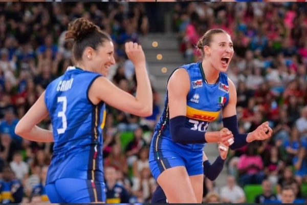 Two female volleyball players in blue uniforms celebrate a point on the court as the crowd cheers in the stands.