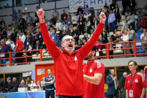 Man in a red jacket raises both arms in triumph amid a cheering crowd at an indoor sports arena.