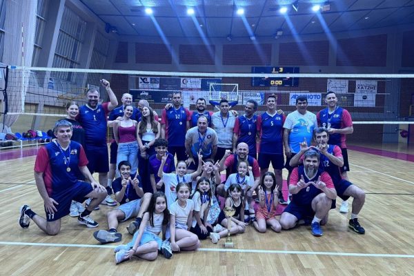 Group photo of a volleyball team (adults and kids) on an indoor court, wearing medals and posing with a trophy after a tournament.