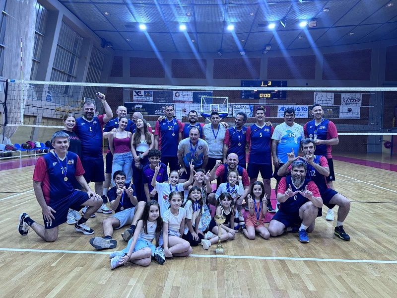 TMEAthliton26k The Volley Lovers Group photo of a volleyball team (adults and kids) on an indoor court, wearing medals and posing with a trophy after a tournament.