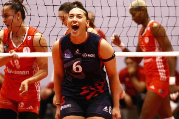 Female volleyball player in a navy uniform with number 6 shouting during a match, net in the foreground, teammates in red behind.