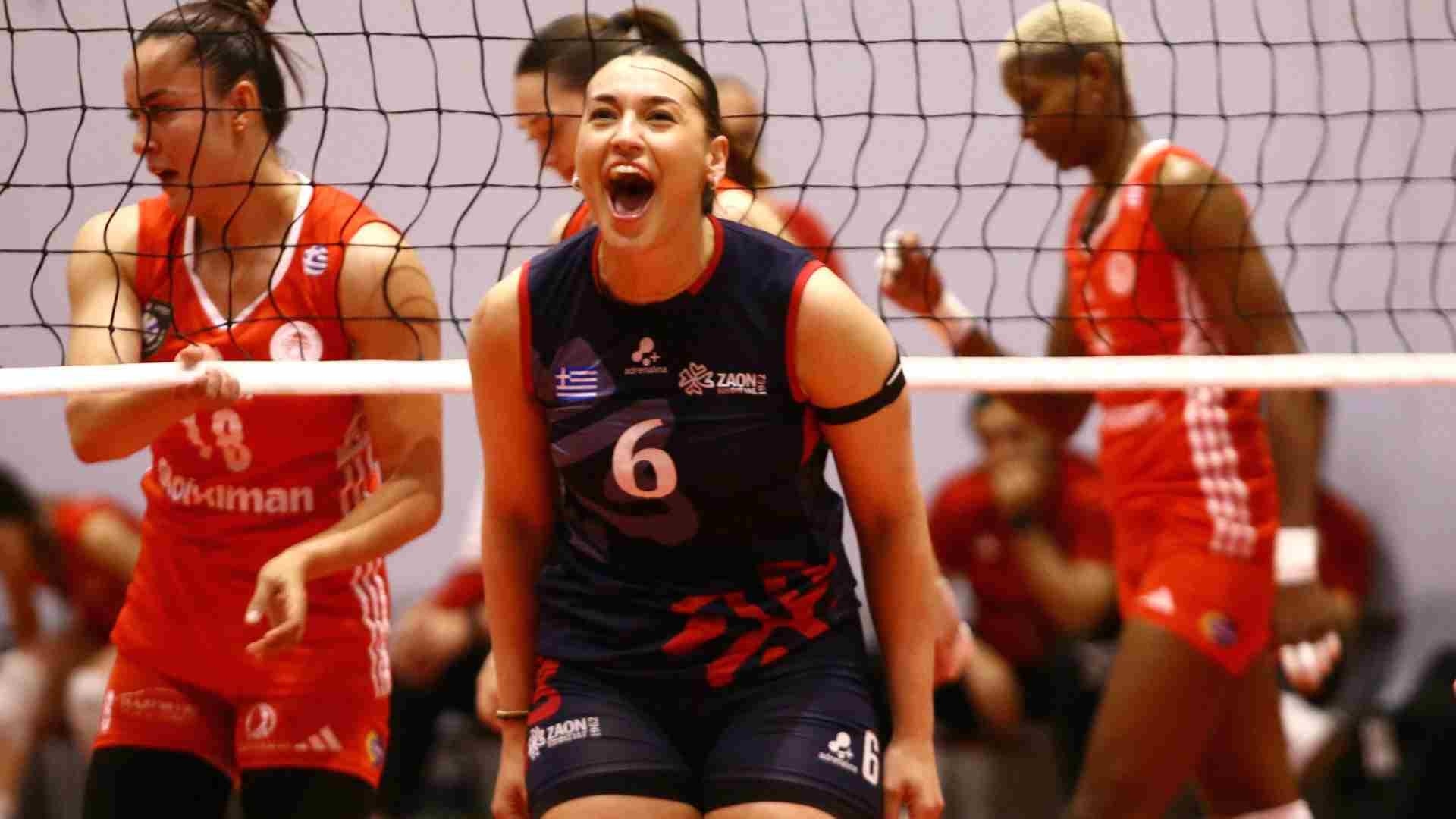 Female volleyball player in a navy uniform with number 6 shouting during a match, net in the foreground, teammates in red behind.