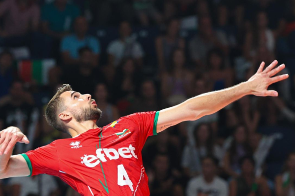 Volleyball player in a red jersey reaches upward with outstretched arms during a game, with a crowded audience in the background.