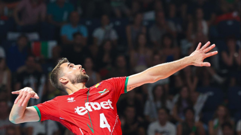 Volleyball player in a red jersey reaches upward with outstretched arms during a game, with a crowded audience in the background.