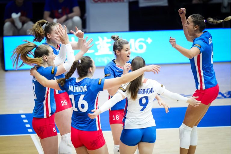 Women’s volleyball team in blue and white uniforms celebrate on the court with raised arms and smiles after a point.