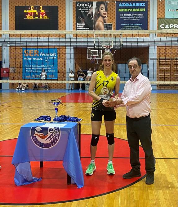 Volleyball player in yellow jersey #17 receives a trophy from a man in a pink shirt on a gym floor with a table of medals beside them.
