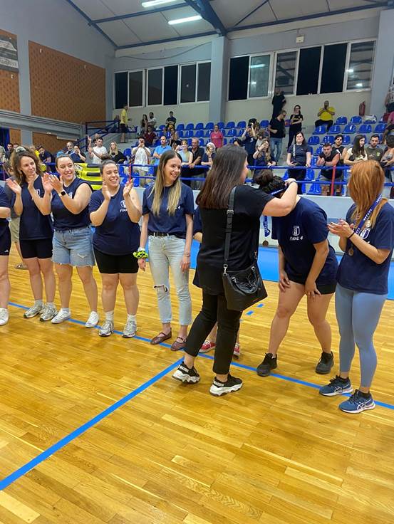 Group of young people in navy shirts line up on a gymnasium floor, clapping, with spectators in blue-seated stands behind them.