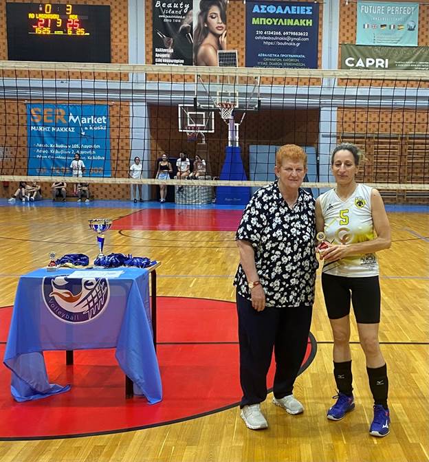 Two women stand on a gym floor after a volleyball award ceremony; the player in a white jersey with number 5 holds a trophy beside a casually dressed woman.