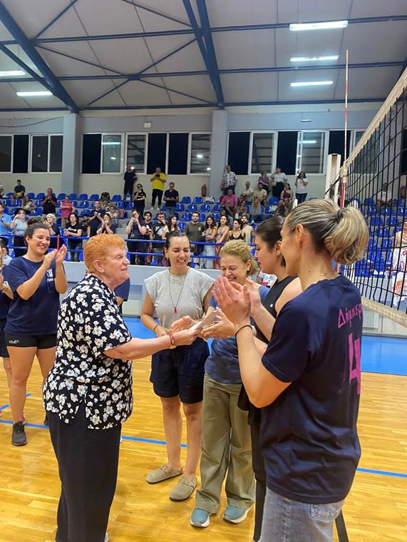 Older woman in a floral shirt hands an item to a volleyball player as teammates applaud on an indoor court, with spectators in the stands.