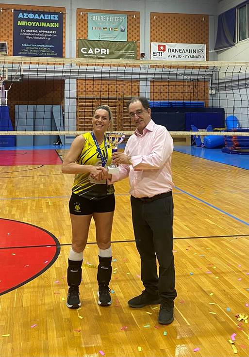 Volleyball player in a yellow jersey accepts a trophy from a man in a pink shirt on an indoor court, net and confetti in the background.