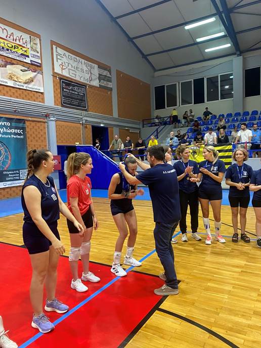 Coach hands out medals to a volleyball player while teammates line up on the court in a gymnasium. Loyal expressions and applause from teammates nearby.