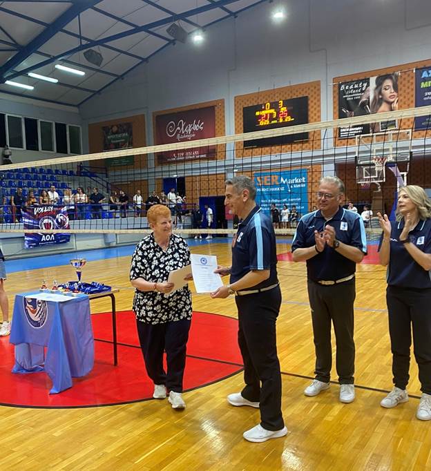 Woman handing a certificate to a man on a volleyball court during an award ceremony, trophies on a table nearby and spectators in the stands behind them.